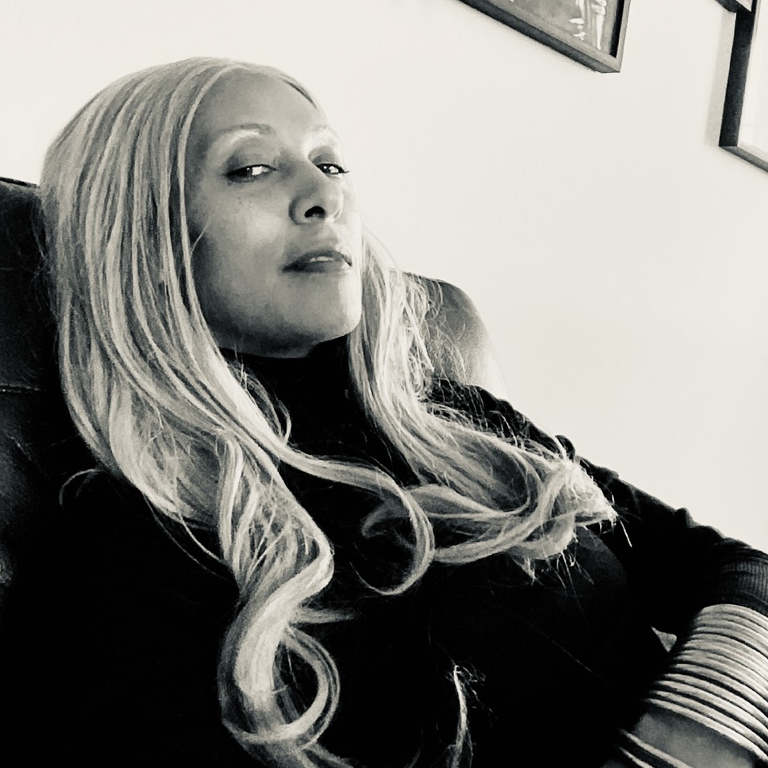 Portrait of Maxximillian Skelevaggio, seated and confident, wearing long light hair and dark clothing.